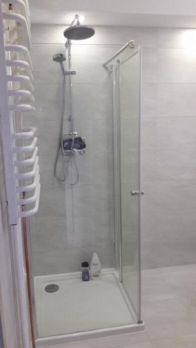 Nowoczesna kabina prysznicowa z deszczownicą i szklanymi drzwiami, widoczny biały grzejnik drabinkowy i dwie butelki kosmetyków na brodziku.