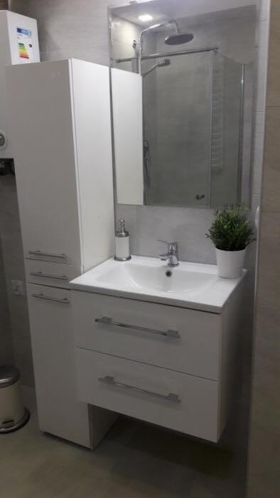 Nowoczesna, biała szafka łazienkowa z umywalką i armaturą, uzupełniona o lustro z kabiną prysznicową w tle oraz dekoracyjną roślinę doniczkową.