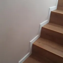 Drewniane schody z białymi listwami przypodłogowymi, przylegające do jasnej, beżowej ściany. Widoczne trzy stopnie z wyraźnym usłojeniem drewna.