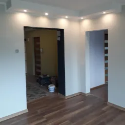 Wnętrze mieszkania w trakcie remontu, widoczne świeżo pomalowane ściany w kolorze białym, drewnopodobne panele na podłodze, oraz narzędzia malarskie i wiadro z farbą w tle.