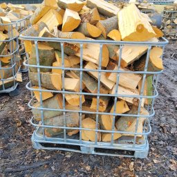 P.U.H. Ewa Milczek - Metalowy kosz wypełniony pociętym drewnem kominkowym na zewnątrz, z budynkiem w tle. Widoczne inne kosze z drewnem. Sezon jesienno-zimowy.