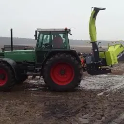 Zielony traktor z podłączoną rębarką do gałęzi na błotnistym polu, w tle widoczne sterty gałęzi i drzew.