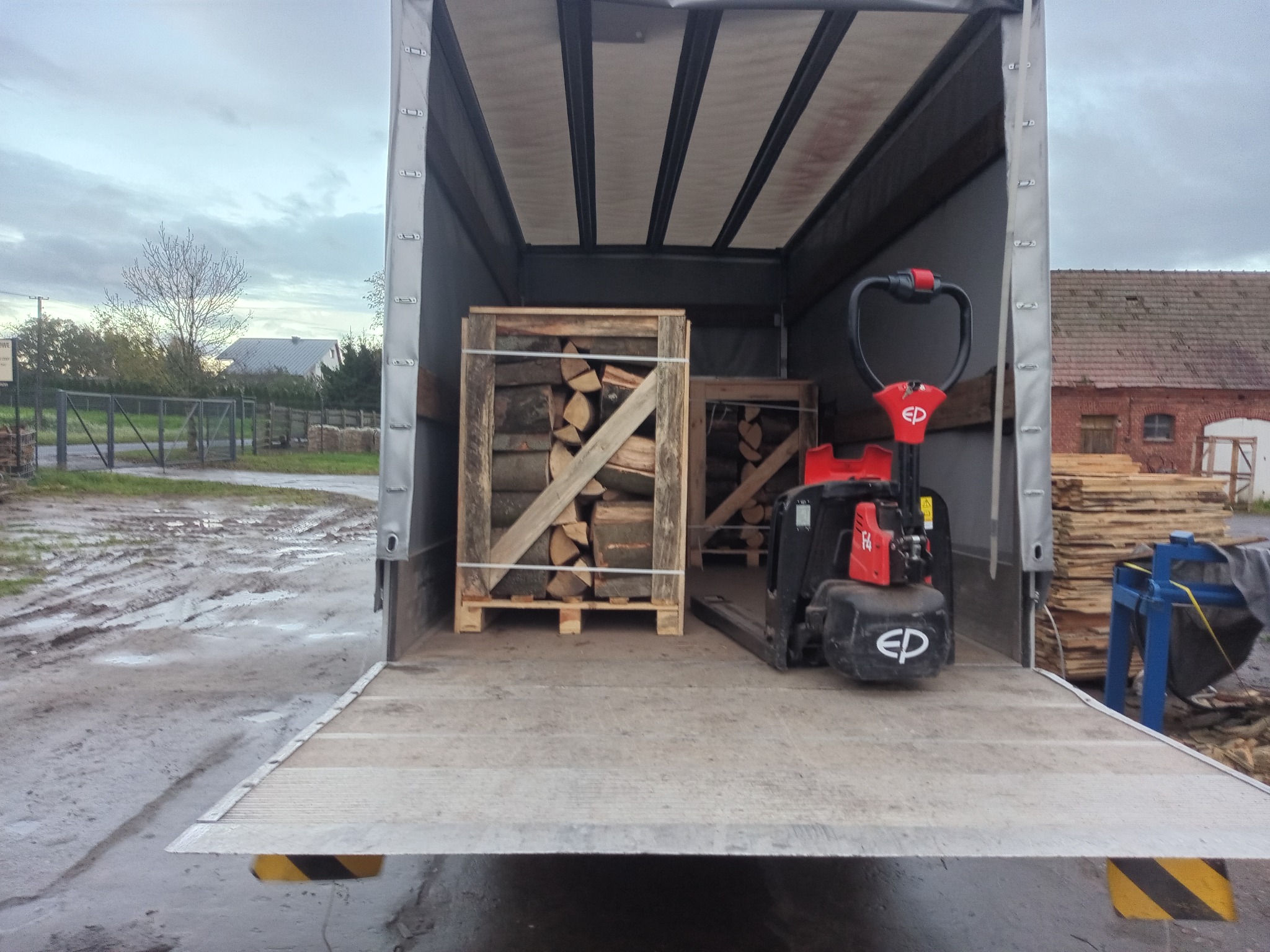 Załadunek drewna kominkowego w skrzyniach na paletach do wnętrza samochodu dostawczego z użyciem wózka paletowego. Widoczne ułożone deski i budynek w tle.