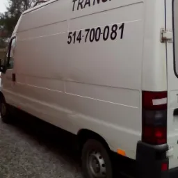 Biały samochód dostawczy z napisem 'TRANSPORT' i numerem telefonu 514-700-081 na boku, zaparkowany na żwirowym podłożu, z widocznymi drzewami w tle.