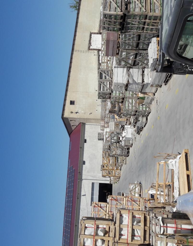 Magazyn z paletami i drewnianymi skrzyniami zawierającymi materiały budowlane, widoczny budynek z panelami słonecznymi na dachu i wózek widłowy.