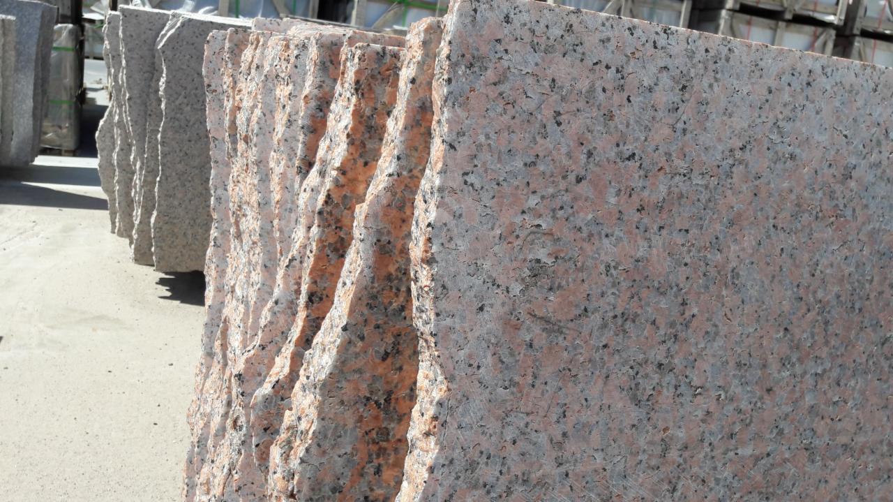 Stos płyt z różowego granitu o szorstkich krawędziach, ułożonych pionowo na zewnątrz, z widocznymi szarymi płytami w tle.