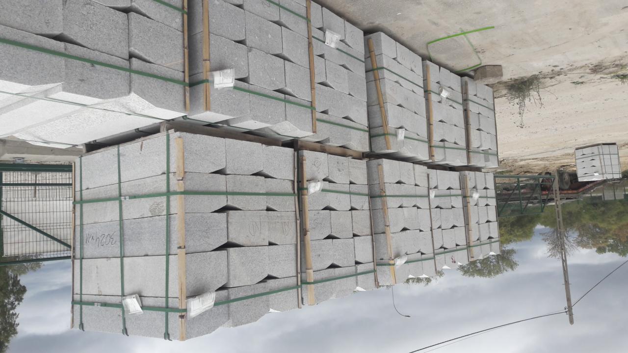 Sztaple szarej kostki brukowej ułożone na paletach, zabezpieczone zieloną taśmą, z widocznymi etykietami i drobnymi napisami na powierzchni kamieni.