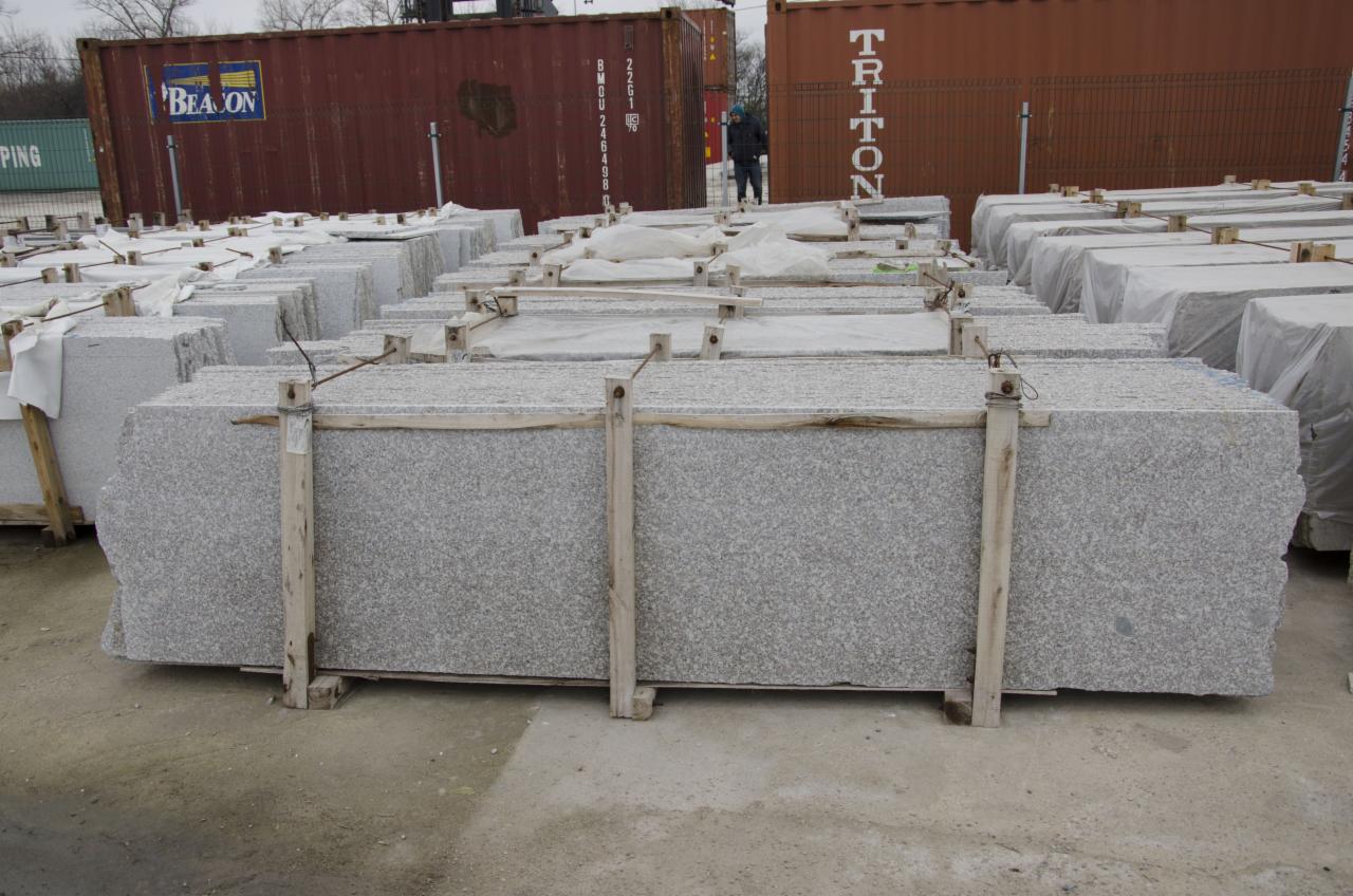 Sztaple szarych płyt kamiennych zabezpieczonych drewnianymi belkami, składowane na placu z widocznymi kontenerami w tle.