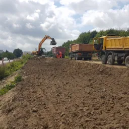 Żółta koparka nabiera ziemię do pomarańczowej wywrotki na placu budowy drogi, obok stoją dwie kolejne ciężarówki, w tle widać ruch samochodów na szosie i zieleń drzew.