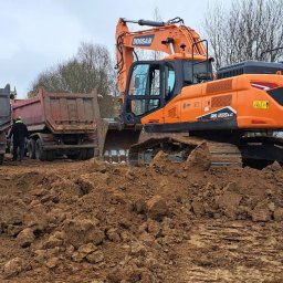 FIRMA USŁUGOWO-TRANSPORTOWA GRE-CAR ŁATA GRZEGORZ - Pomarańczowa koparka Doosan i dwie ciężarówki na placu budowy z wykopaną ziemią. W tle budynki mieszkalne i drzewa.
