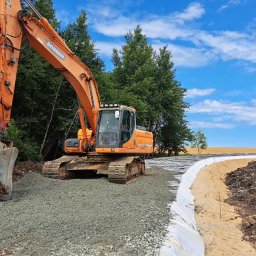 FIRMA USŁUGOWO-TRANSPORTOWA GRE-CAR ŁATA GRZEGORZ - Pomarańczowa koparka Doosan na placu budowy, przygotowuje podłoże pod drogę. Widoczne warstwy gruntu i geowłóknina. Błękitne niebo z chmurami w tle.
