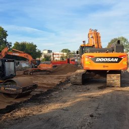 FIRMA USŁUGOWO-TRANSPORTOWA GRE-CAR ŁATA GRZEGORZ - Żółty walec JCB i pomarańczowe koparki Doosan na placu budowy w słoneczny dzień. W tle budynki i drzewa. Widoczny znak Agencji Ochrony Szabel.