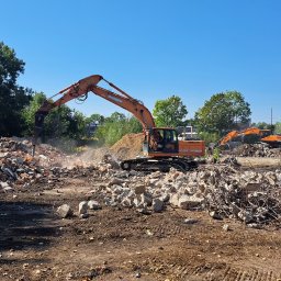 FUT GRE-CAR Łata Grzegorz - Pomarańczowy koparko-młot hydrauliczny kruszy gruz na placu budowy. W tle widoczna kolejna maszyna i zieleń. Błękitne niebo.