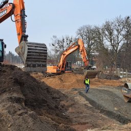 FIRMA USŁUGOWO-TRANSPORTOWA GRE-CAR ŁATA GRZEGORZ - Dwie pomarańczowe koparki Doosan i żółty walec JCB na placu budowy. Pracownik w żółtej kamizelce przechadza się po terenie. W tle drzewa i elementy infrastruktury.