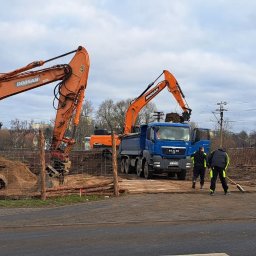 FIRMA USŁUGOWO-TRANSPORTOWA GRE-CAR ŁATA GRZEGORZ - Pomarańczowe koparki Doosan i niebieska ciężarówka MAN na placu budowy. Dwóch robotników z miotłami. W tle budynki mieszkalne i pochmurne niebo.