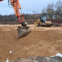 FIRMA USŁUGOWO-TRANSPORTOWA GRE-CAR ŁATA GRZEGORZ - Pomarańczowa koparka z łyżką i walec drogowy na placu budowy z piaskiem. W tle widoczne drzewa i budynki.