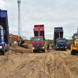 FIRMA USŁUGOWO-TRANSPORTOWA GRE-CAR ŁATA GRZEGORZ - Ciężarówki wyładowują piasek na placu budowy, w tle koparka i ubijarka. Widoczne ślady opon na piasku i pracownik w kamizelce odblaskowej.