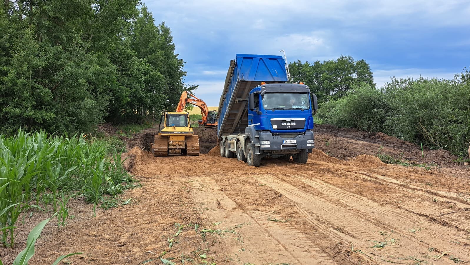 Niebieska wywrotka MAN wysypuje ziemię na placu budowy, w tle żółty spychacz Caterpillar i koparka Hitachi, teren przygotowywany pod budowę, widok z przodu.