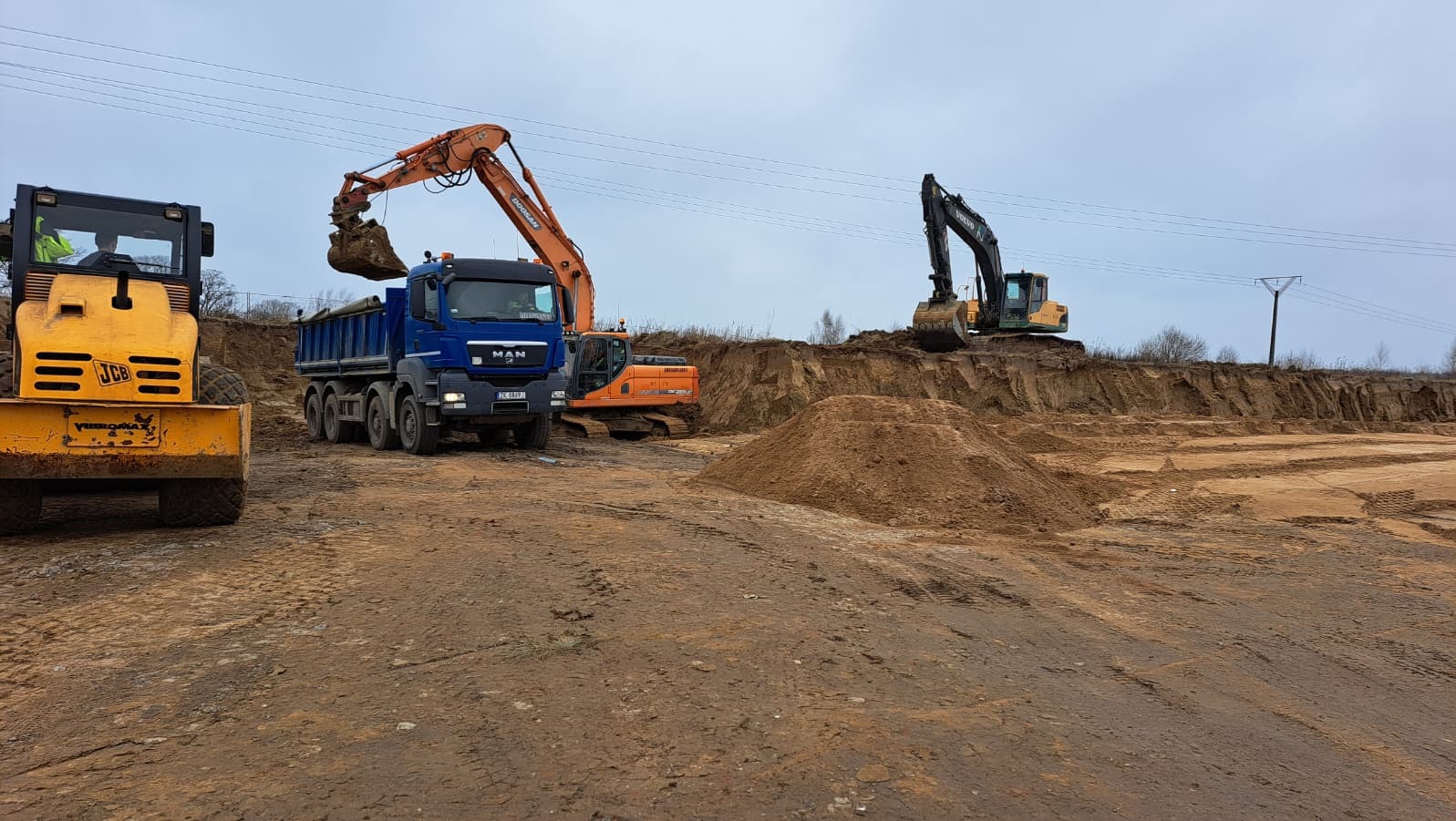 Żółty walec, niebieska ciężarówka MAN i pomarańczowa koparka Doosan w trakcie prac ziemnych. W tle koparka Volvo na skarpie. Prace ziemne w toku.