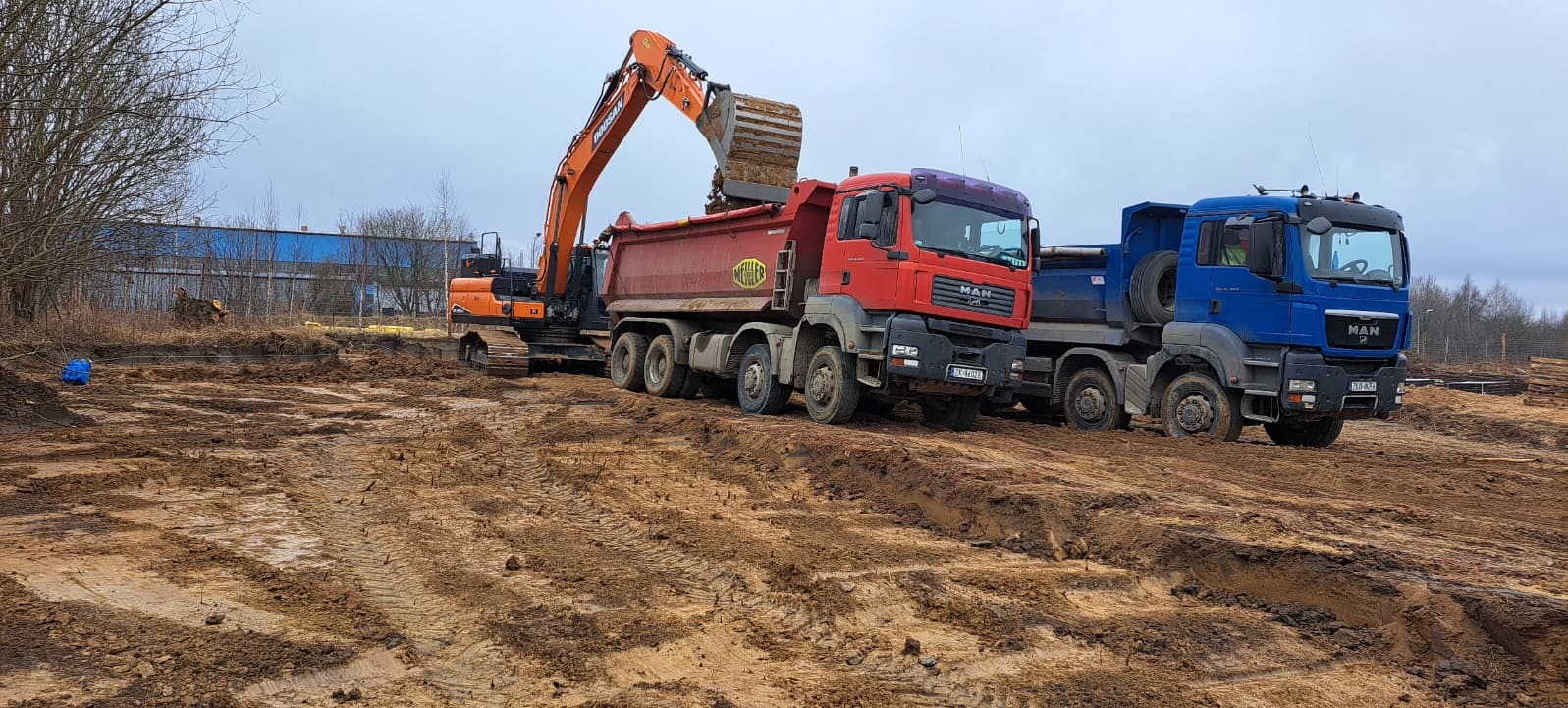 Pomarańczowa koparka ładuje ziemię do czerwonej ciężarówki MAN na placu budowy. W tle błękitna ciężarówka i budynek. Widoczne ślady opon na błotnistym terenie.