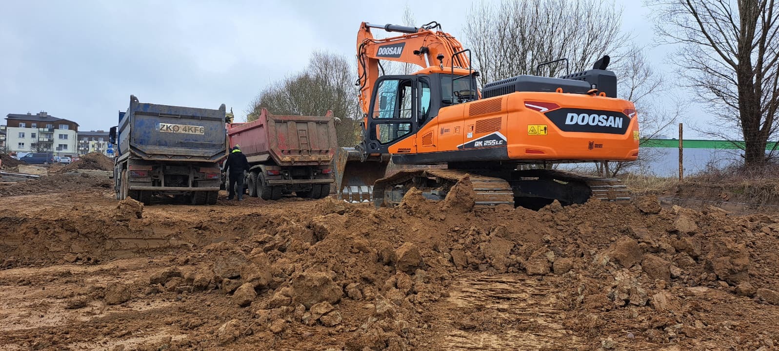 Pomarańczowa koparka Doosan i dwie ciężarówki na placu budowy z wykopaną ziemią. W tle budynki mieszkalne i drzewa.