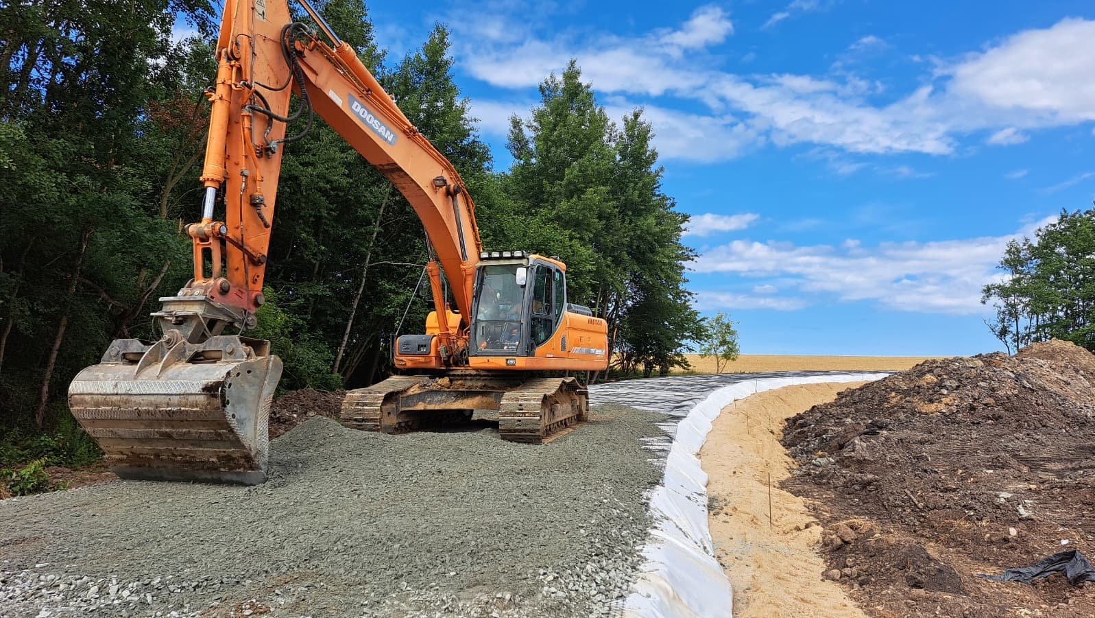 Pomarańczowa koparka Doosan na placu budowy, przygotowuje podłoże pod drogę. Widoczne warstwy gruntu i geowłóknina. Błękitne niebo z chmurami w tle.