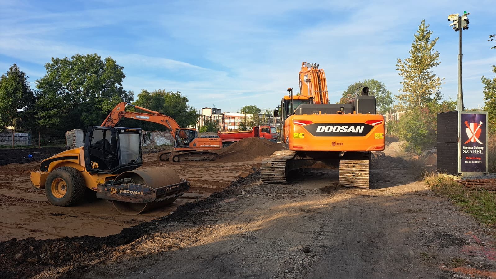 Żółty walec JCB i pomarańczowe koparki Doosan na placu budowy w słoneczny dzień. W tle budynki i drzewa. Widoczny znak Agencji Ochrony Szabel.