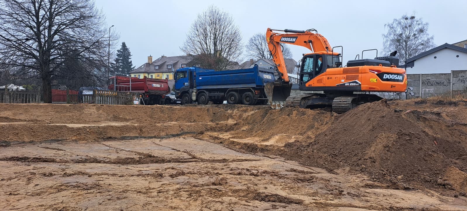 Pomarańczowa koparka Doosan i dwie ciężarówki na placu budowy. Widoczne ślady pracy koparki w ziemi. W tle budynki mieszkalne i drzewa bez liści.