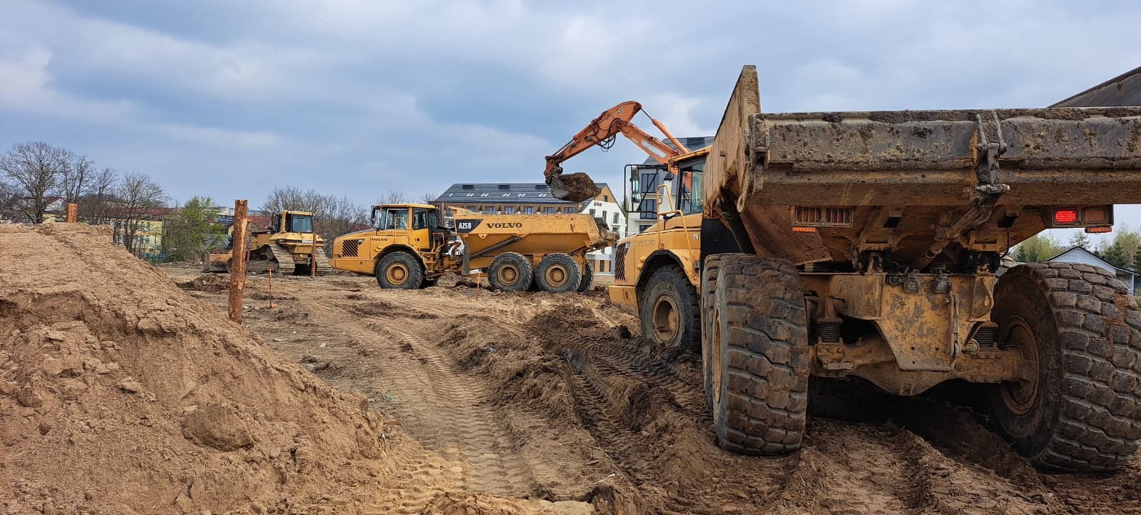 Żółty wywrotka Volvo A25D w trakcie załadunku ziemi przez koparkę. W tle widoczne inne maszyny budowlane i budynki.
