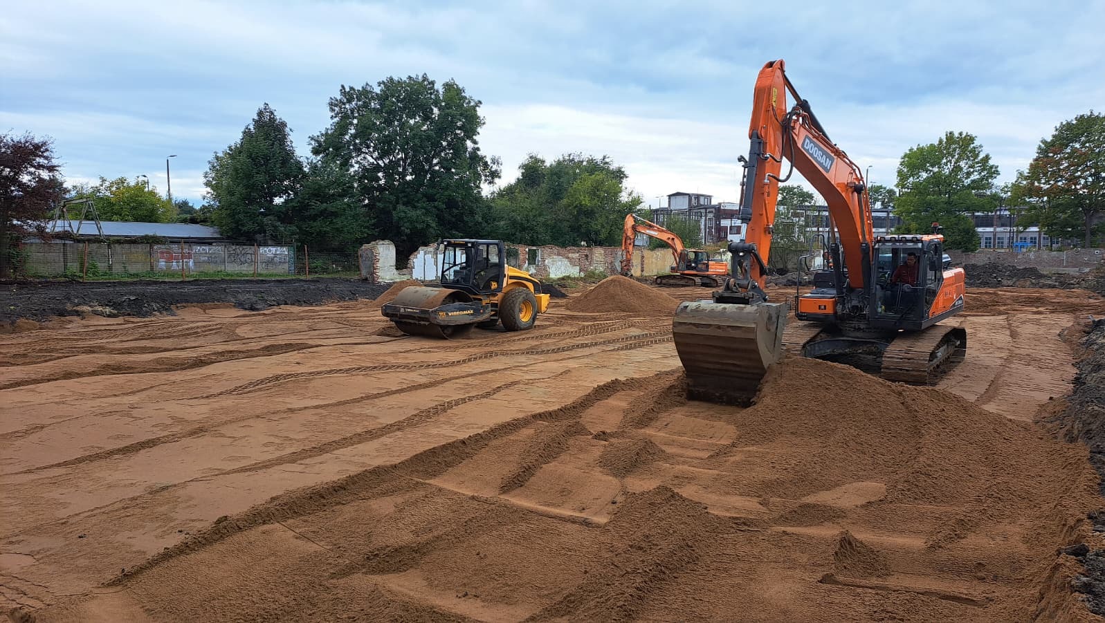 Niwelacja terenu z użyciem walca i koparek Doosan. Widoczne ślady opon na wyrównanym, piaszczystym podłożu. W tle zarośla i fragmenty zburzonych budynków.