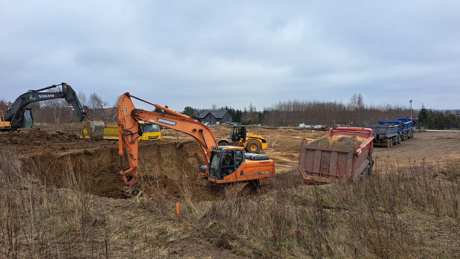 Praca koparki Doosan przy wykopie w Koszalinie. W tle widoczne ciężarówki i inny sprzęt budowlany. Pochmurne niebo.