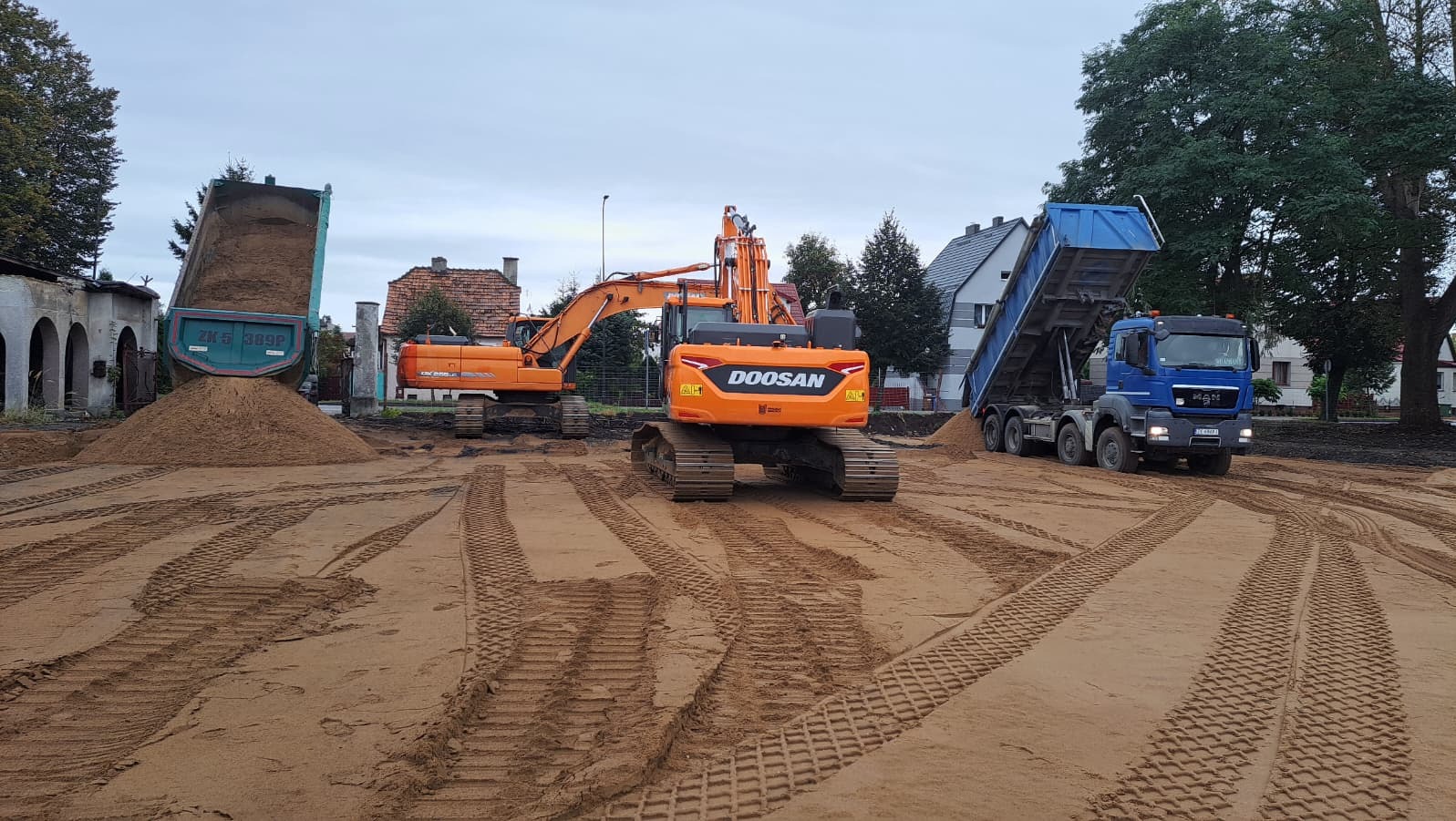 Dwa wywrotki i koparka Doosan na placu budowy. Jeden wywrotka wysypuje piasek. Ślady opon na piasku. W tle budynki mieszkalne i drzewa.