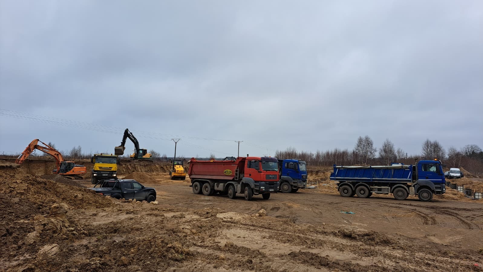 Praca wre: Dwa różne modele koparek, walec i kilka ciężarówek na placu budowy, w tle pochmurne niebo. Widoczne ślady opon na ziemi.