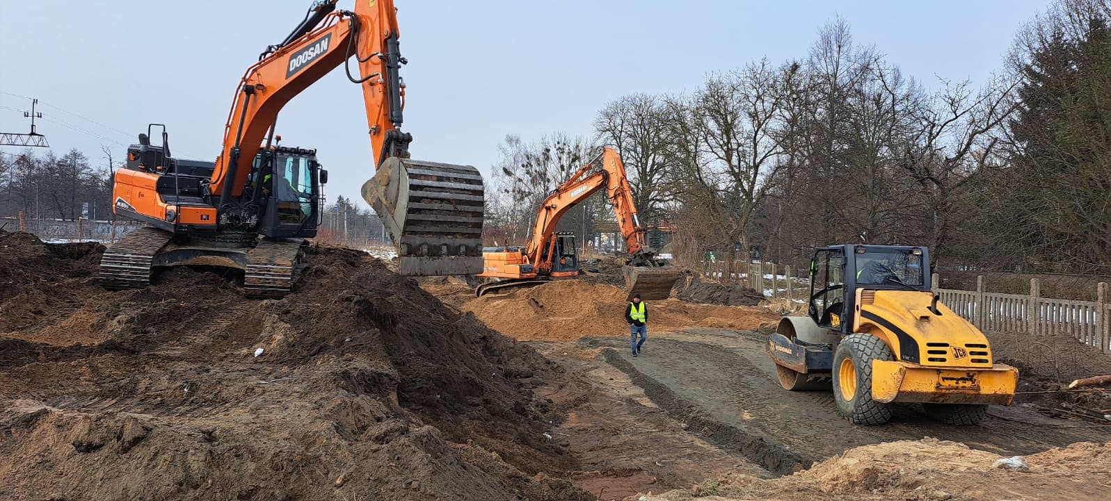 Dwie pomarańczowe koparki Doosan i żółty walec JCB na placu budowy. Pracownik w żółtej kamizelce przechadza się po terenie. W tle drzewa i elementy infrastruktury.