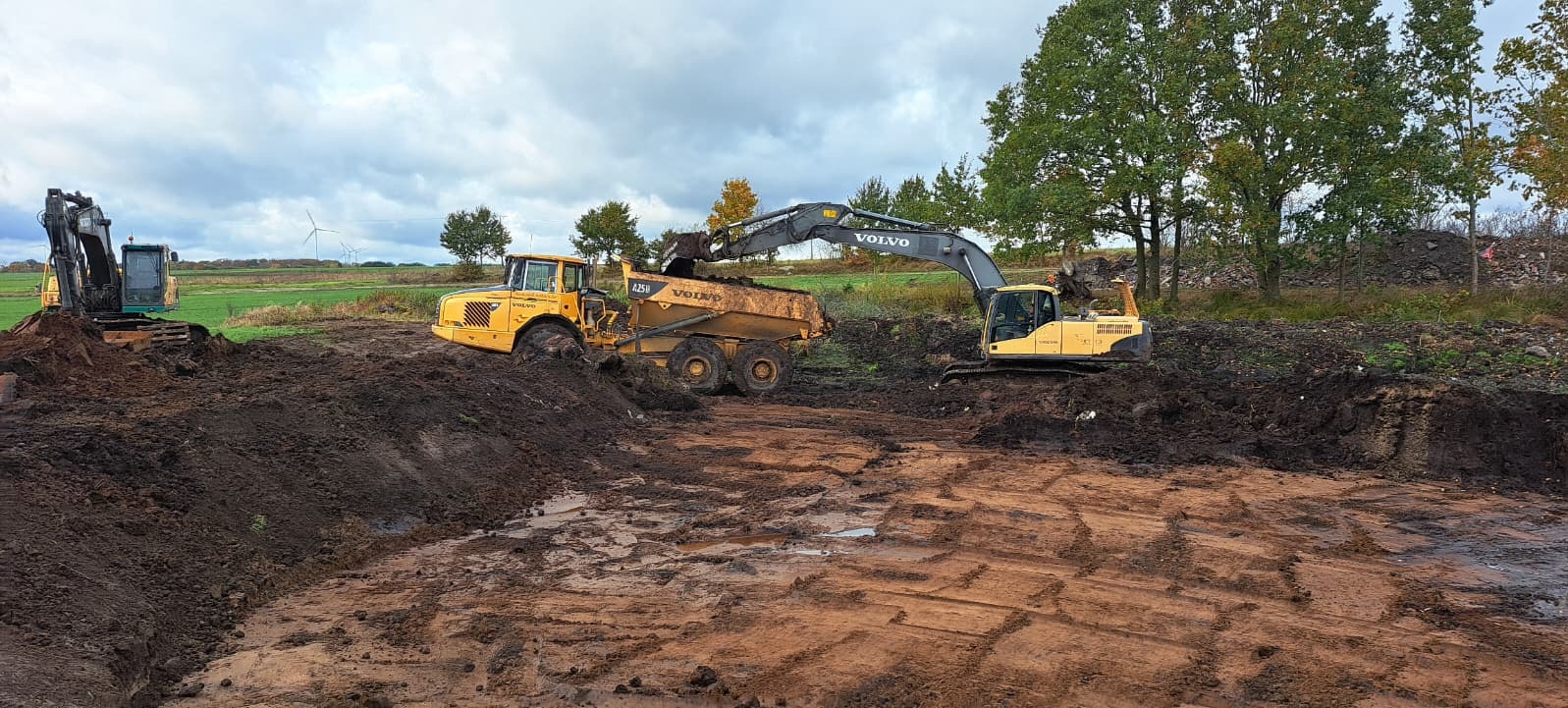 Żółta wywrotka Volvo A25G i koparka Volvo EC220E w trakcie prac ziemnych na polu. W tle widoczne turbiny wiatrowe i drzewa.