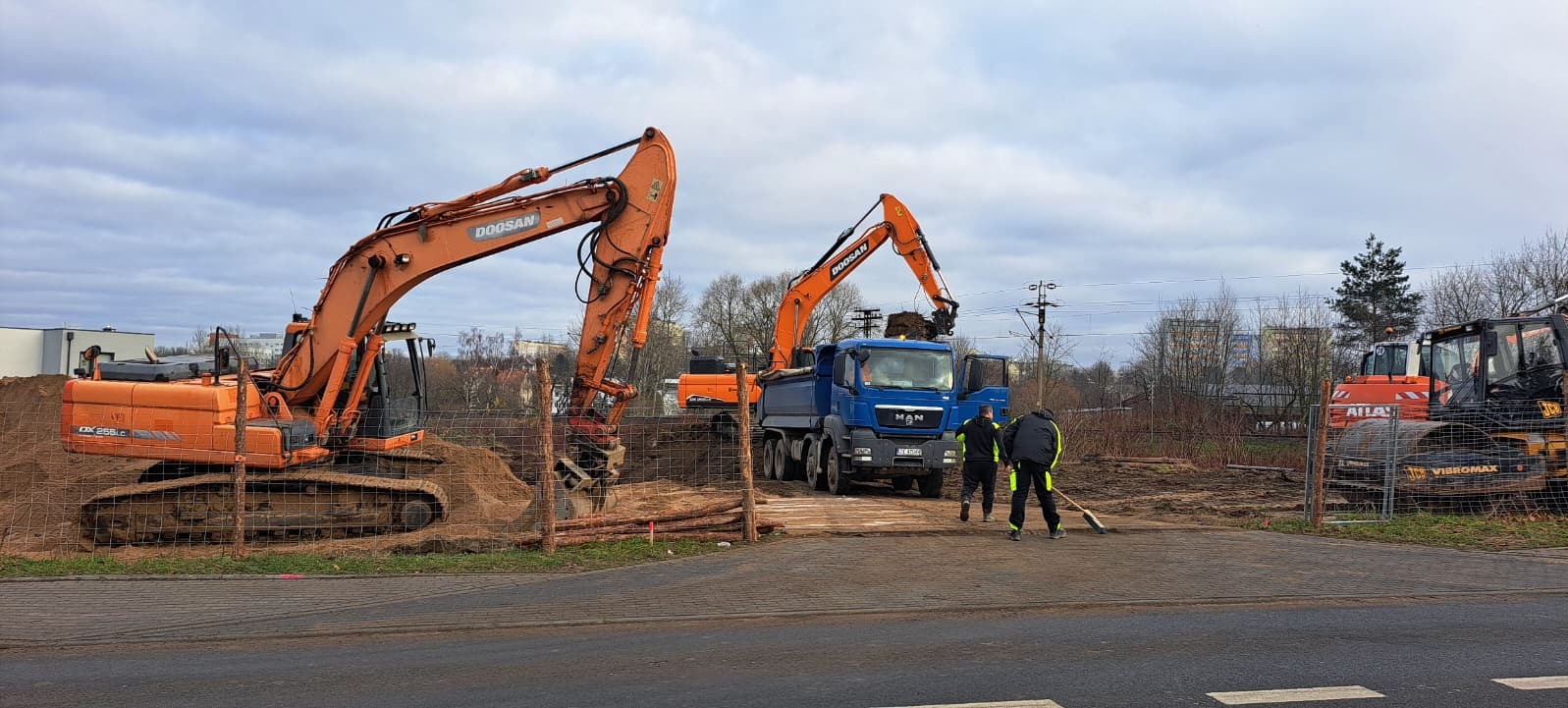 Pomarańczowe koparki Doosan i niebieska ciężarówka MAN na placu budowy. Dwóch robotników z miotłami. W tle budynki mieszkalne i pochmurne niebo.