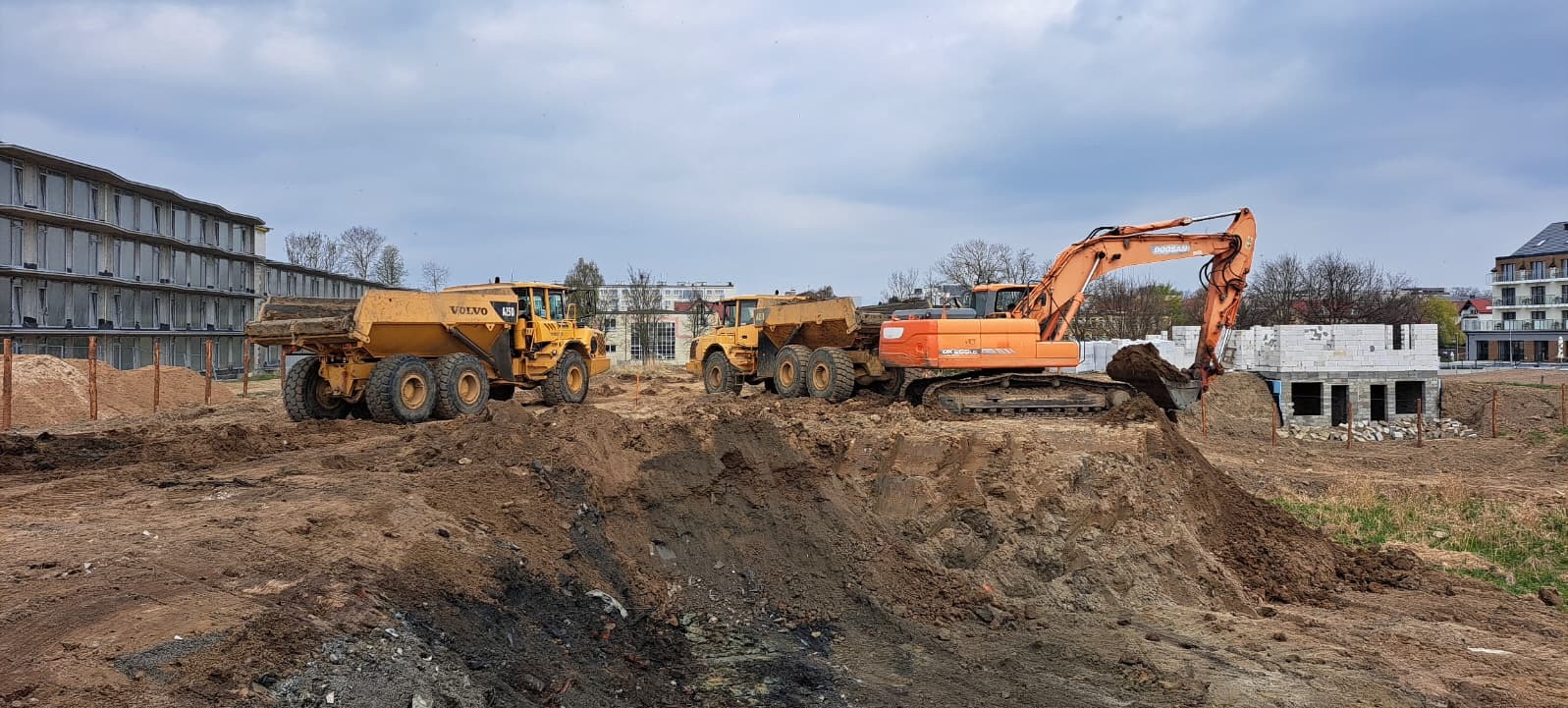 Żółte wywrotki Volvo i pomarańczowa koparka Doosan na placu budowy z fundamentami budynku w tle, w pochmurny dzień.