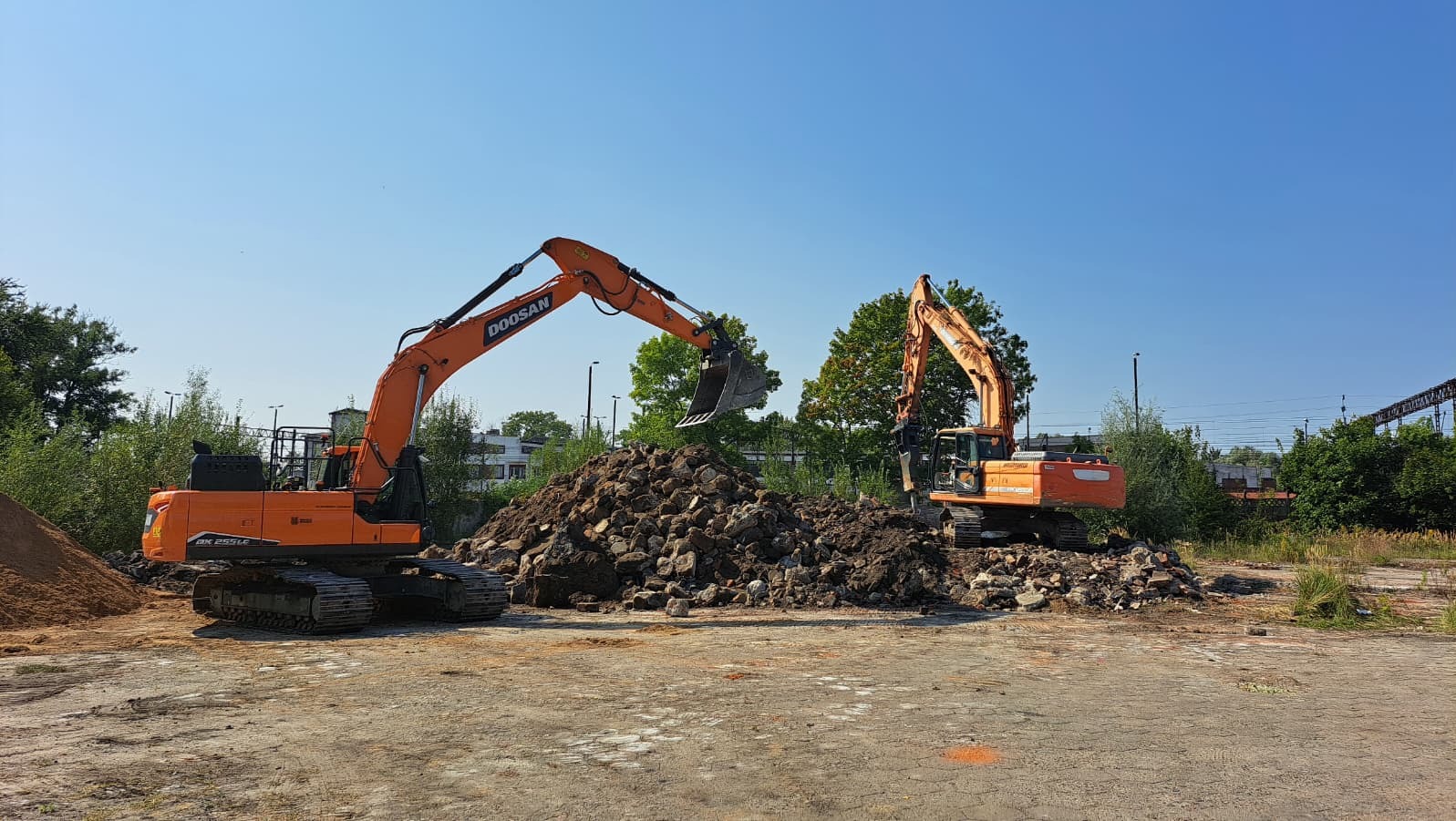 Dwie pomarańczowe koparki Doosan pracują na placu budowy, usypując górę gruzu i ziemi na tle błękitnego nieba i zieleni. Widoczne elementy infrastruktury miejskiej.