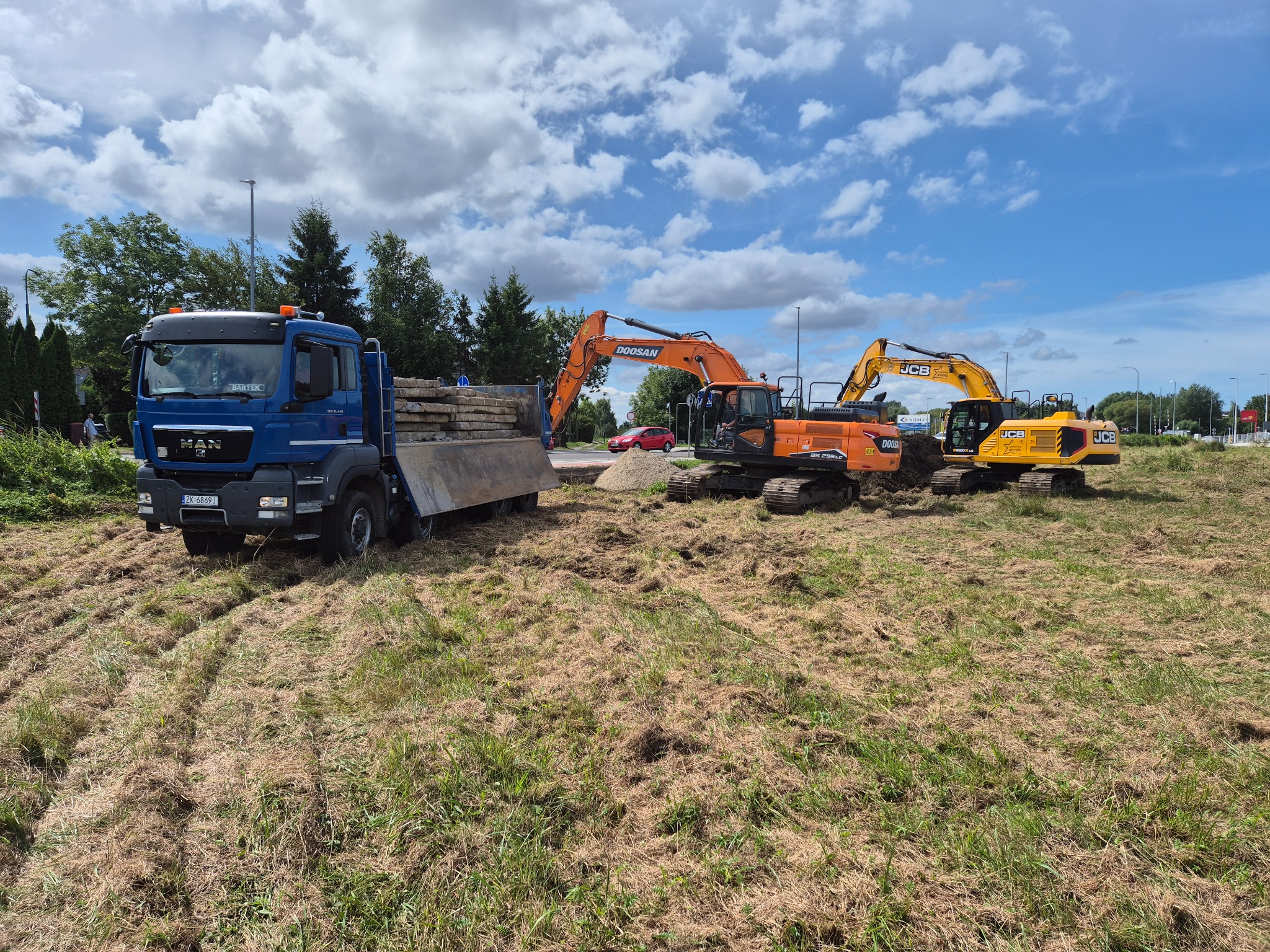 Ciężarówka MAN z ładunkiem i dwa pomarańczowo-żółte  ekskawatory na polu z częściowo skoszoną trawą, błękitne niebo z chmurami w tle.