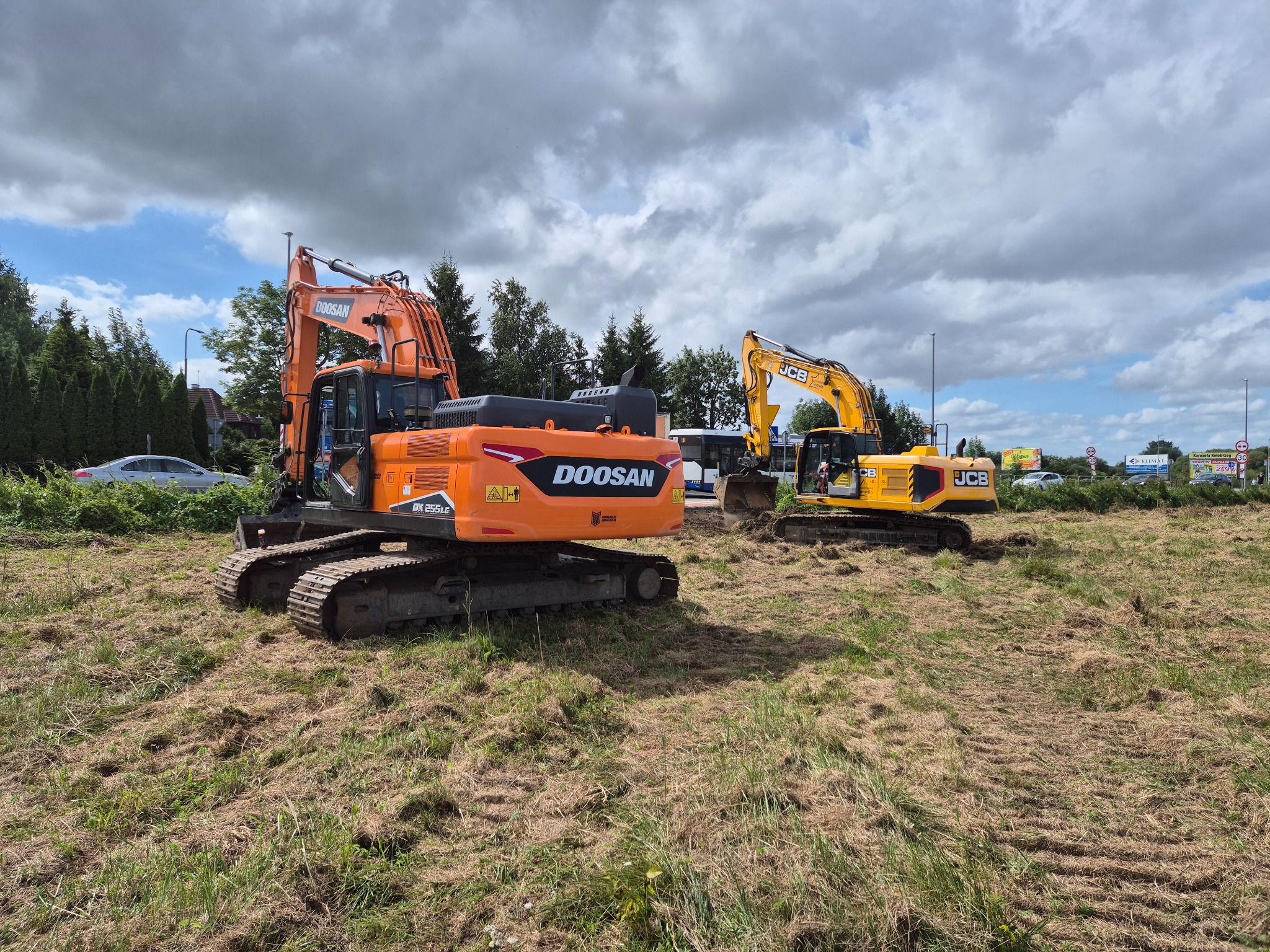 Dwa pracujące na trawiastym terenie: pomarańczowy Doosan i żółty JCB. W tle drzewa i budynki, nad nimi pochmurne niebo. Maszyny w trakcie robót ziemnych.