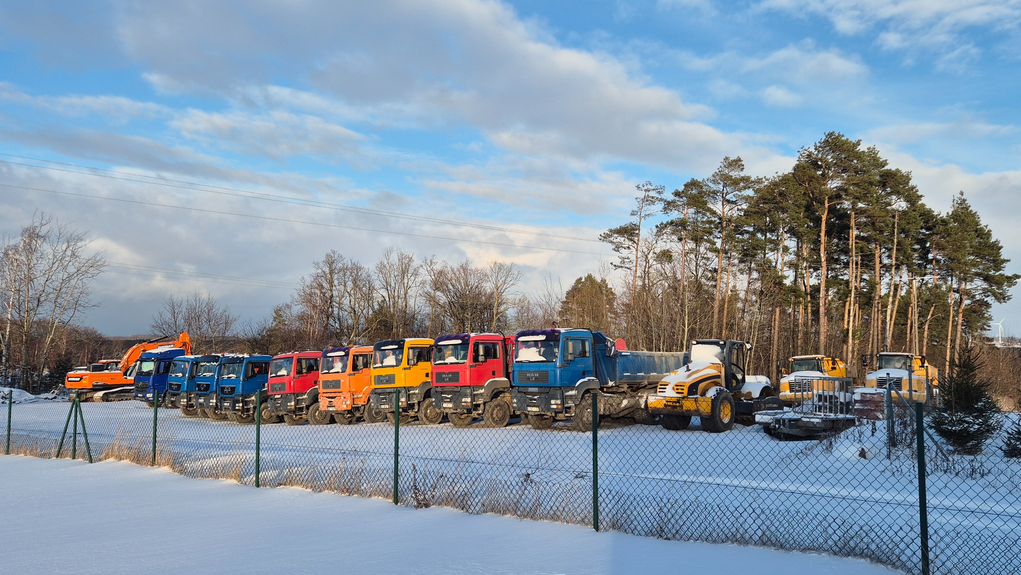 Rząd kolorowych ciężarówek MAN i koparek na zasypanym śniegiem placu budowy, otoczonym siatką ogrodzeniową i drzewami, pod błękitnym niebem z chmurami.