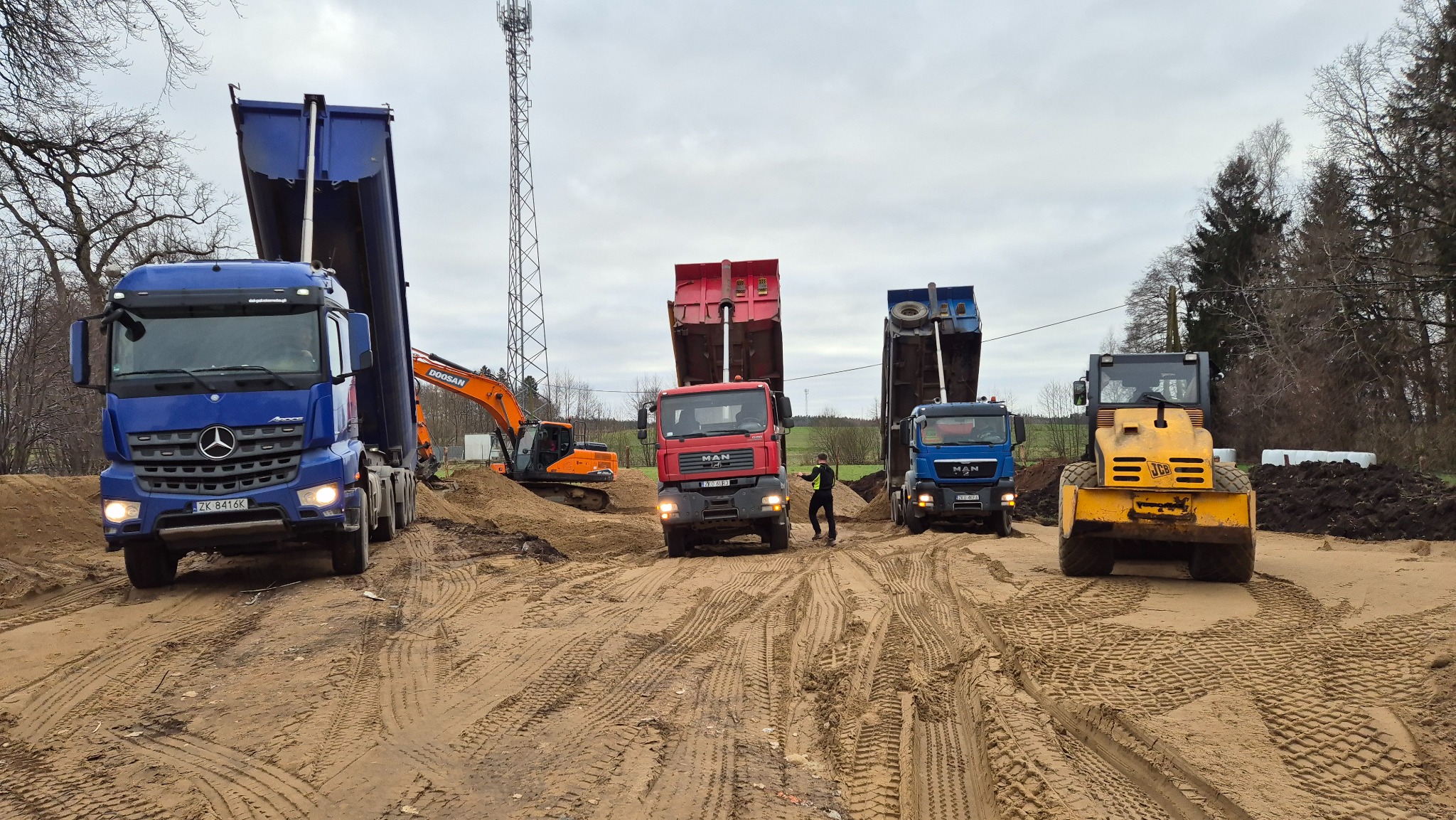Ciężarówki wyładowują piasek na placu budowy, w tle koparka i ubijarka. Widoczne ślady opon na piasku i pracownik w kamizelce odblaskowej.