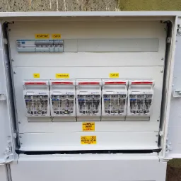 Otwarta, biała skrzynka elektryczna z bezpiecznikami różnicowoprądowymi marki Apator, oznaczonymi etykietami 'Podgrzewacz', 'OSW/GN', 'WC', 'Hydrofornia' i 'Camping', zamontowana na ścianie...