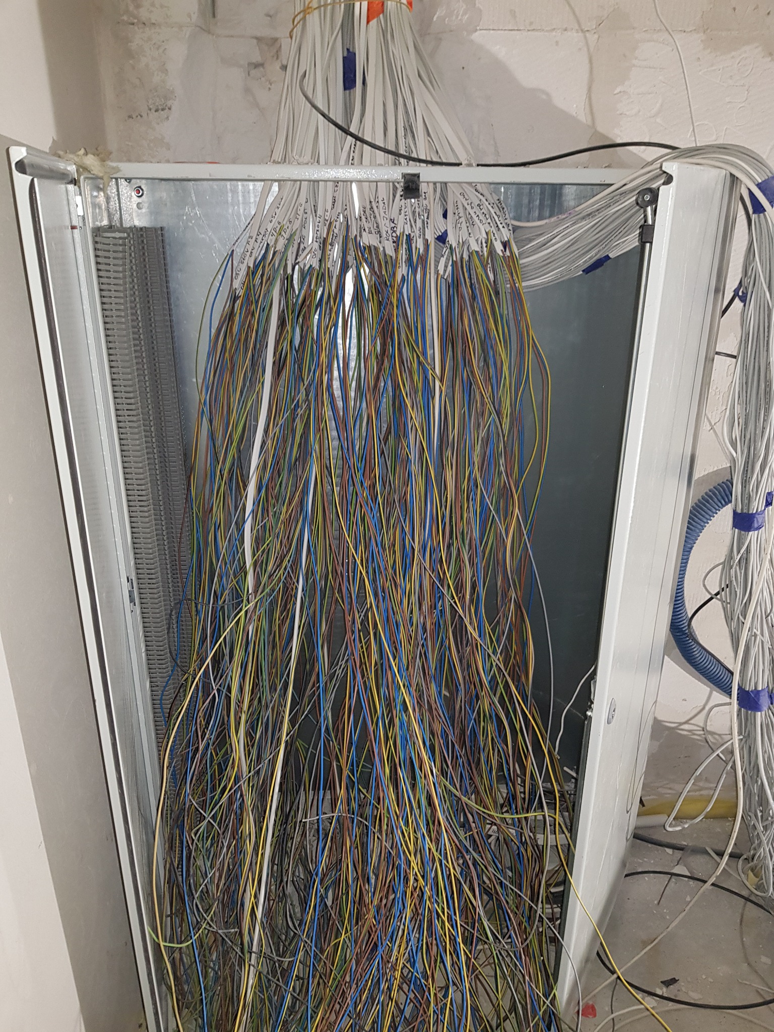 Skomplikowana instalacja elektryczna w trakcie montażu: liczne, kolorowe kable z etykietami zwisają z metalowej obudowy, widoczne elementy montażowe i peszle w tle, surowy stan wykończenia wnętrza.