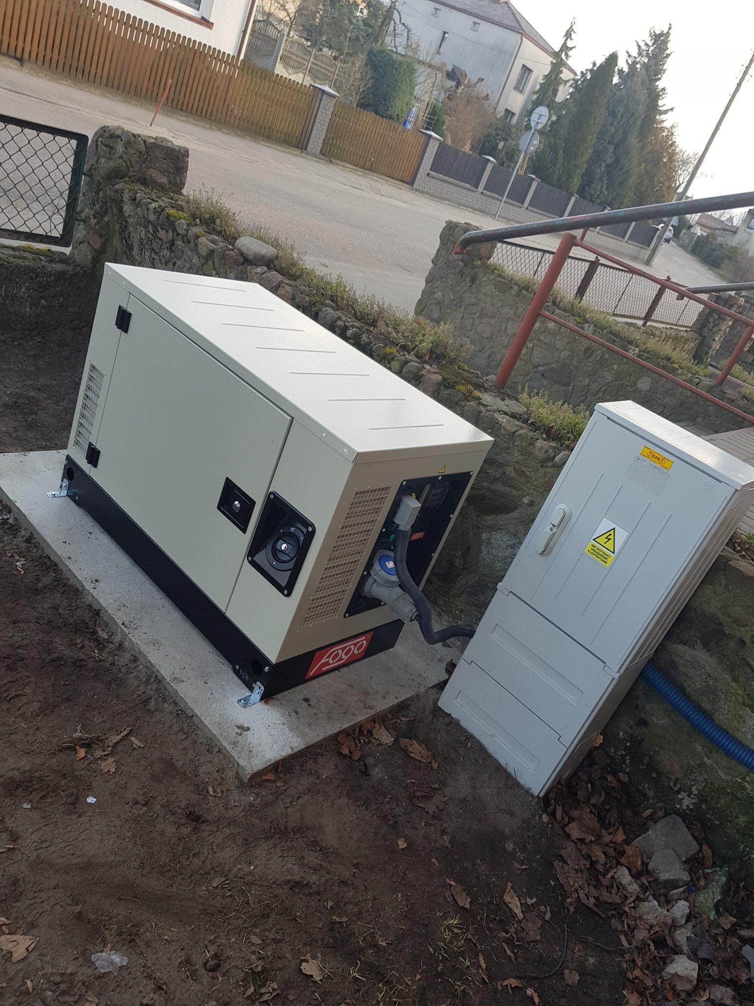 Agregat prądotwórczy zamontowany na betonowym fundamencie obok skrzynki elektrycznej z ostrzeżeniem o wysokim napięciu, na zewnątrz w dzień.