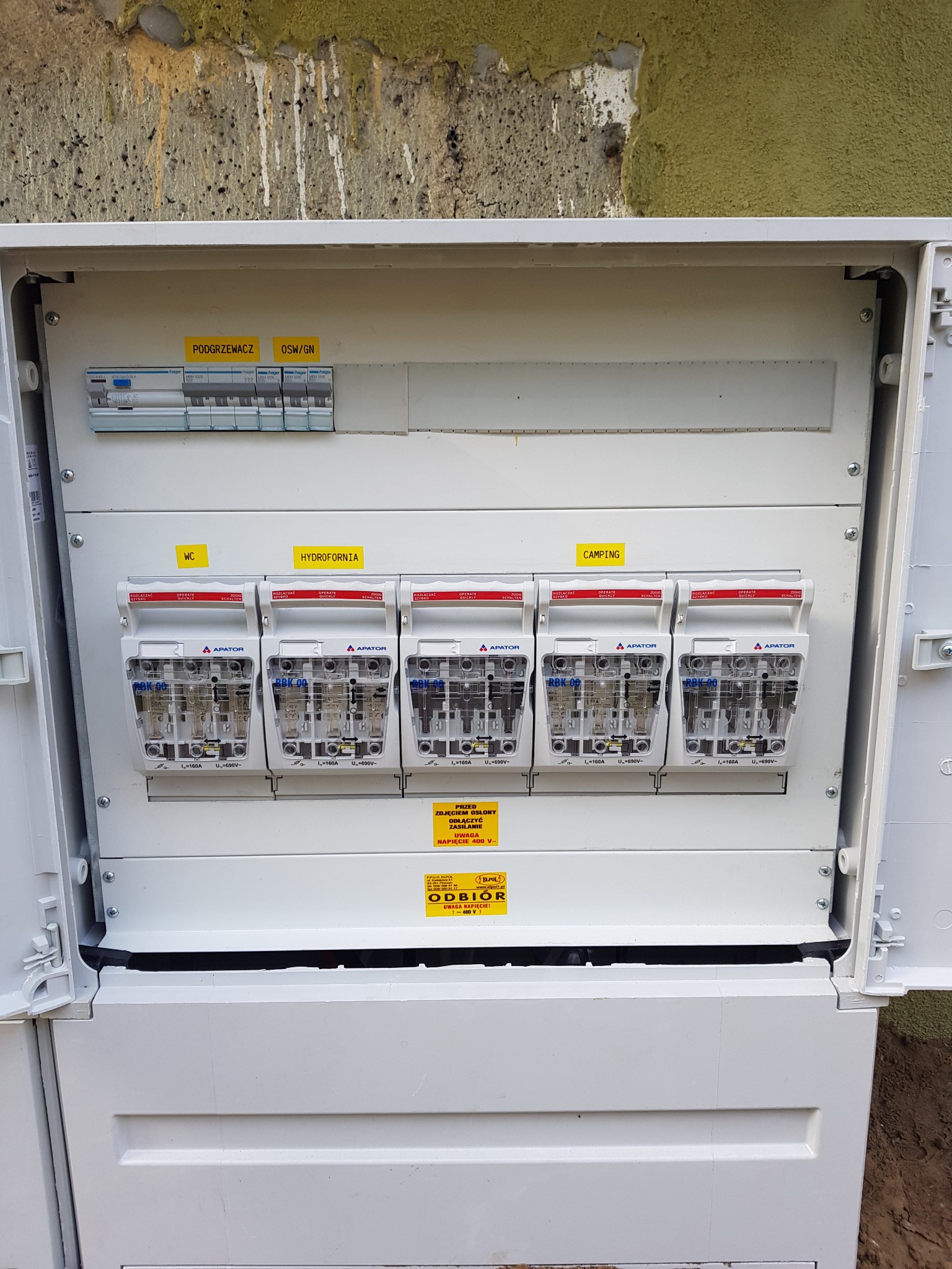 Otwarta, biała skrzynka elektryczna z bezpiecznikami różnicowoprądowymi marki Apator, oznaczonymi etykietami 'Podgrzewacz', 'OSW/GN', 'WC', 'Hydrofornia' i 'Camping', zamontowana na ścianie...