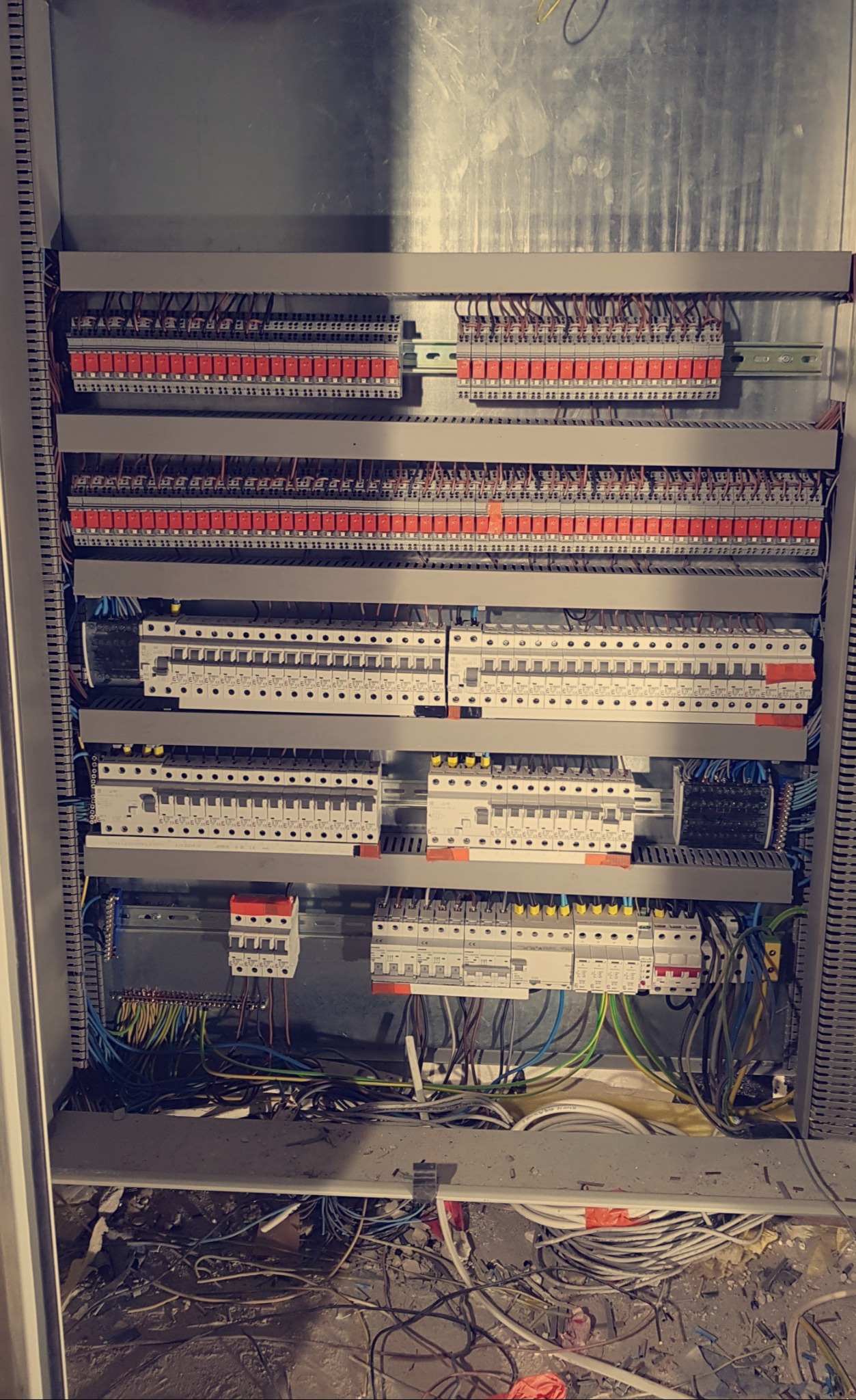 Otwarta szafa elektryczna z rozbudowanym okablowaniem, widoczne liczne bezpieczniki, złączki i przewody różnego koloru, pod spodem gruz i resztki kabli.