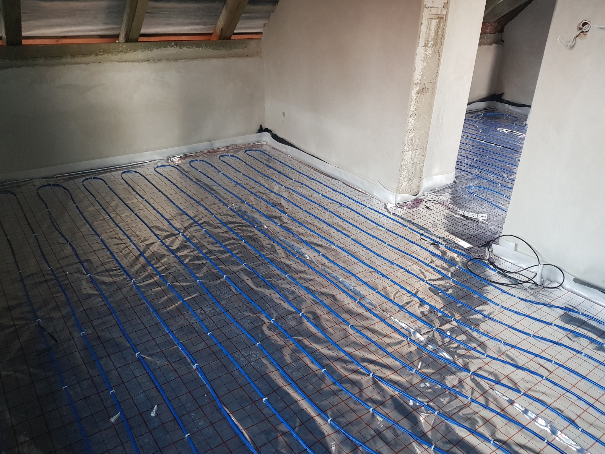 Niedokończona instalacja ogrzewania podłogowego z niebieskimi rurkami na folii aluminiowej w surowym wnętrzu poddasza.