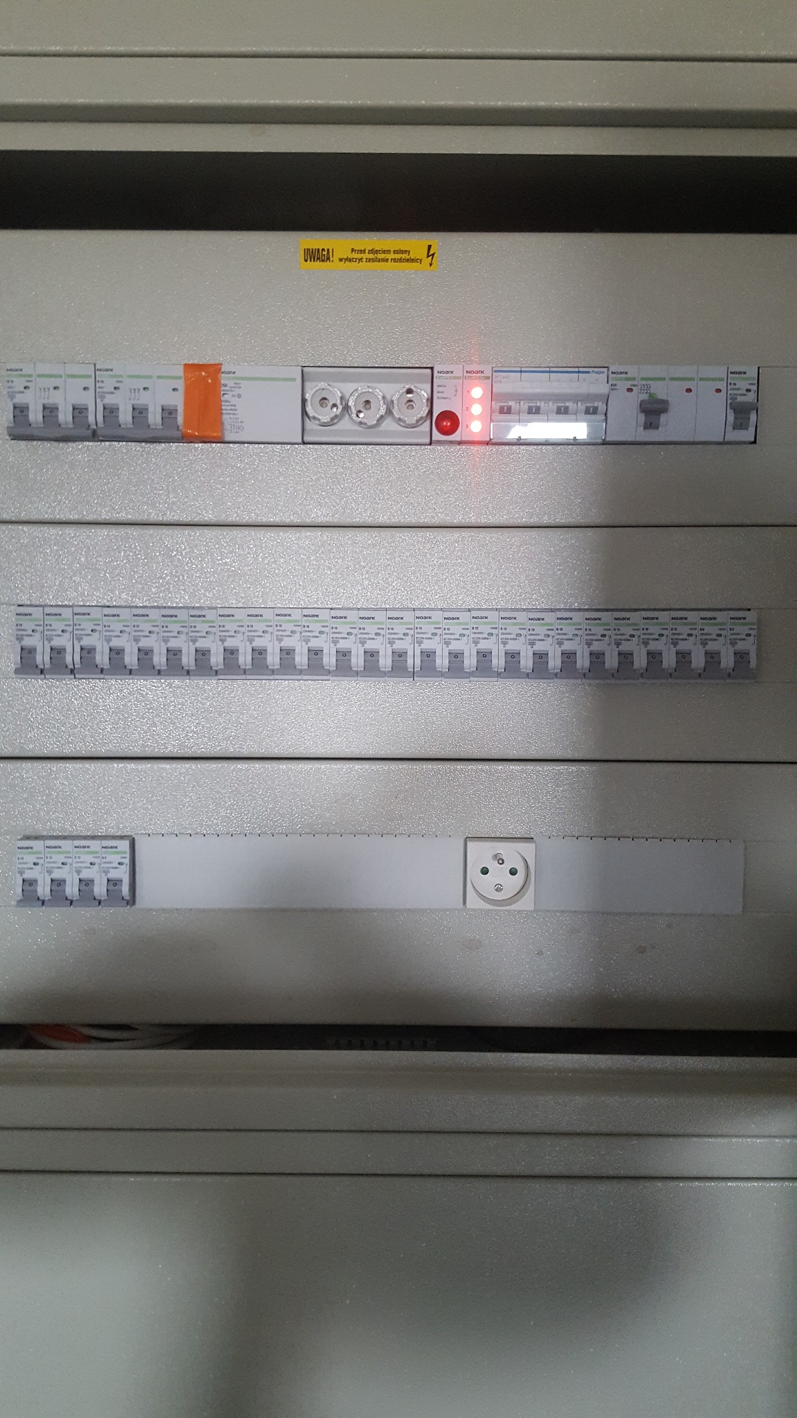 Rozdzielnica elektryczna z bezpiecznikami, wyłącznikami i gniazdem w Lubichowie, widoczne czerwone diody sygnalizacyjne i żółta naklejka ostrzegawcza.
