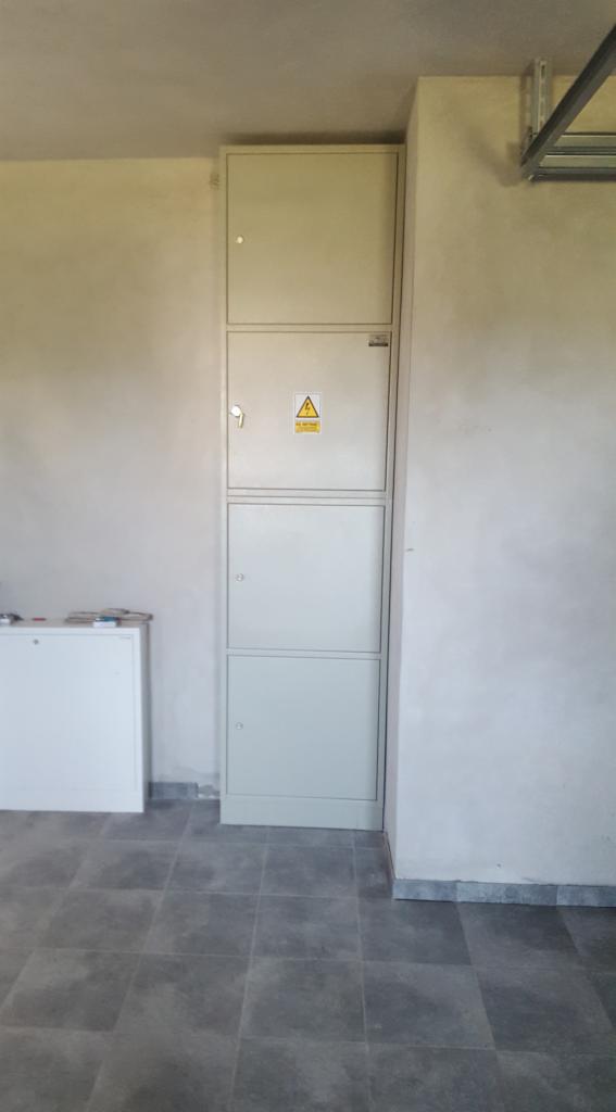 Trzyczęściowa, metalowa szafa elektryczna z żółtym znakiem ostrzegawczym wysokiego napięcia, zamontowana na ścianie w surowym stanie, na szarej podłodze z płytek.